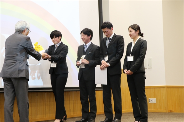 写真：ステージに登壇した学生4名が鵜崎学長から賞状や記念品をもらう様子
