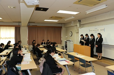 写真：教室のホワイトボードの前に並んで発表する4名の学生と、それを聴講するそれ以外の学生