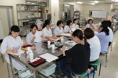 写真：テーブルを囲んで座る学生6名と人見准教授、マルイ担当者4名。学生と人見准教授は白衣を着用している。