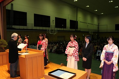 写真：ステージ上に並ぶ7名の受賞者。一人は賞状を読み上げる学長の前に立っている