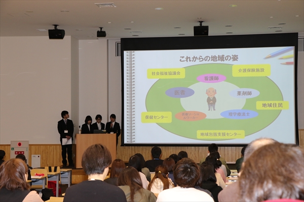写真：大勢の参加者の前で、スライドを使用してプレゼンをする4名の学生