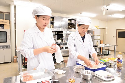 写真：試食を繰り返す学生3人。調理台に材料がいっぱい並ぶ