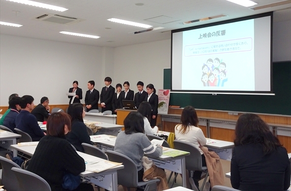 写真：7人の学生がスライドの前に立って取り組みを発表する様子。それを見る他大学の学生たち。