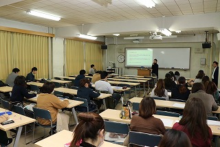 写真：教室の教壇で話す秋山涼子氏と、聴講する学生