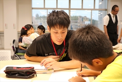 写真：感想文を書く小学生の様子を見ている男子学生