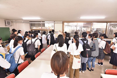写真：校内施設見学で集団給食室を説明する学生スタッフ。それを聞く参加者多数