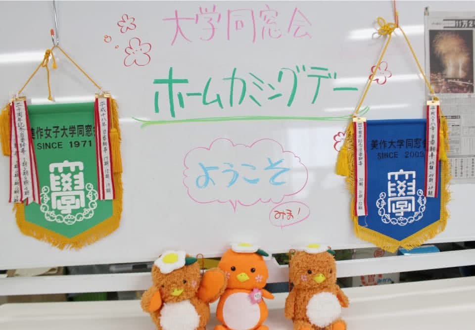写真:ホワイトボードに書かれた「大学同窓会 ホームカミングデー ようこそみま!」という文字と、美作大学同窓会の旗2つと3体のみまっぱちゃんのぬいぐるみ