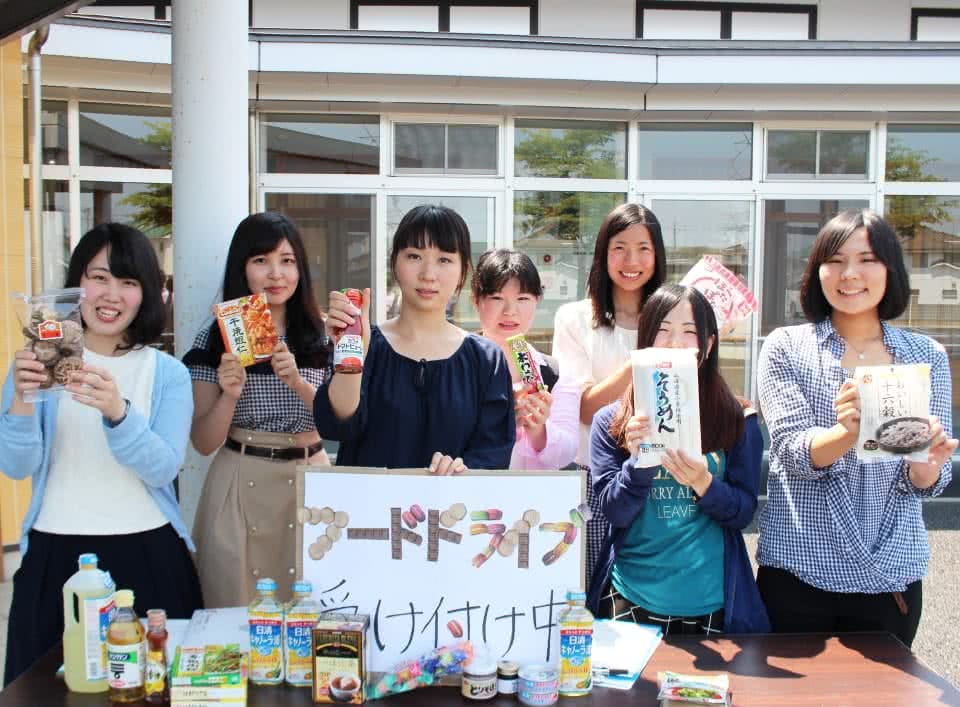 写真:「フードドライブ受け付け中」と書かれたパネルと色々な食品が置かれたテーブルの後ろに、食品を手に持って立つ食品ロス削減サークルのメンバーたち