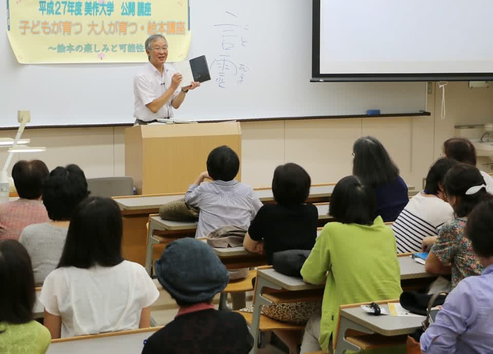 写真:「平成27年度 美作大学 公開講座 子どもが育つ 大人が育つ 絵本講座 ~絵本の楽しみと可能性~」と講座名が書かれた紙が貼られたホワイトボードの前で公開講座を行う講師と、受講する地域の人々。