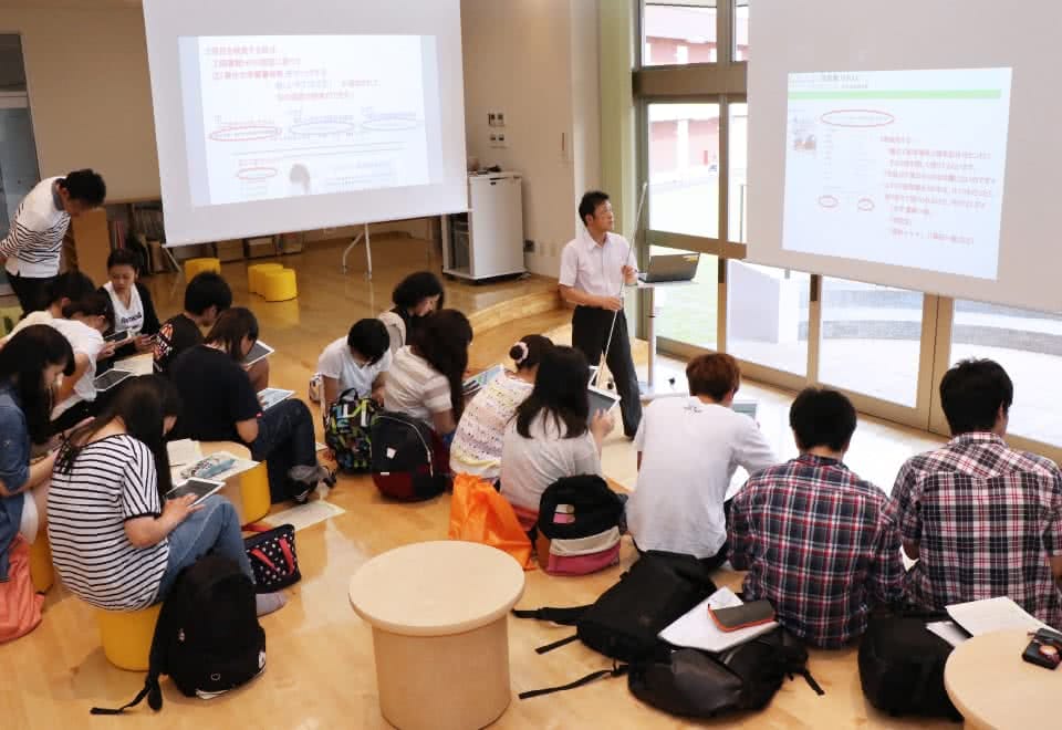 写真:サークルコモンズのステージ中央でプロジェクターを使った授業を行っている教員と、それを聞いている学生たち。