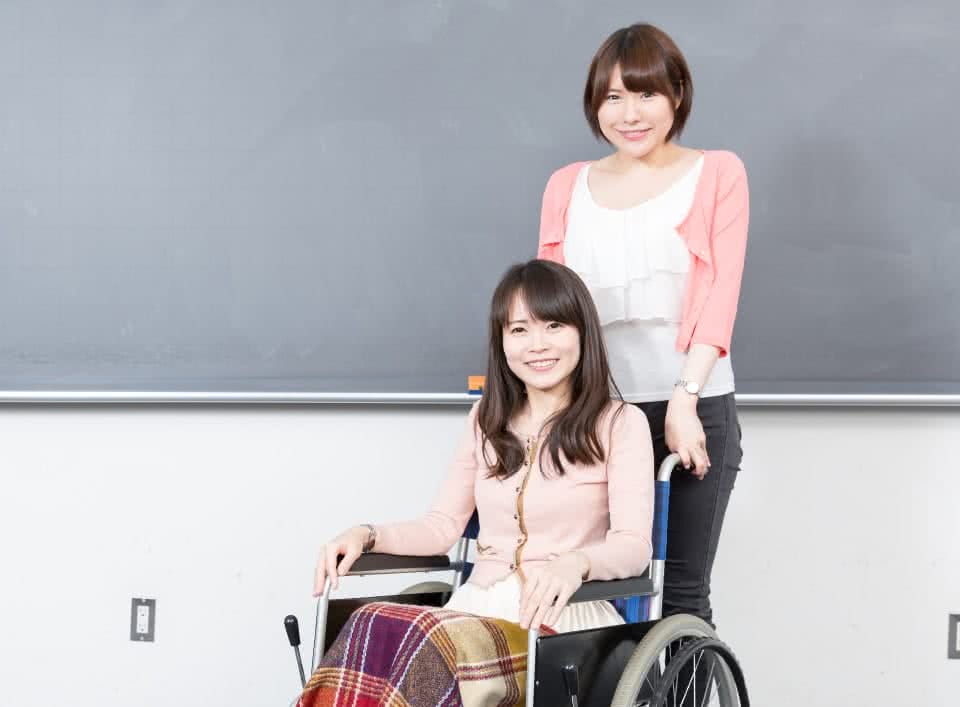 写真:黒板の前で車椅子に座っている女子学生と、その車椅子をもつ女子学生。