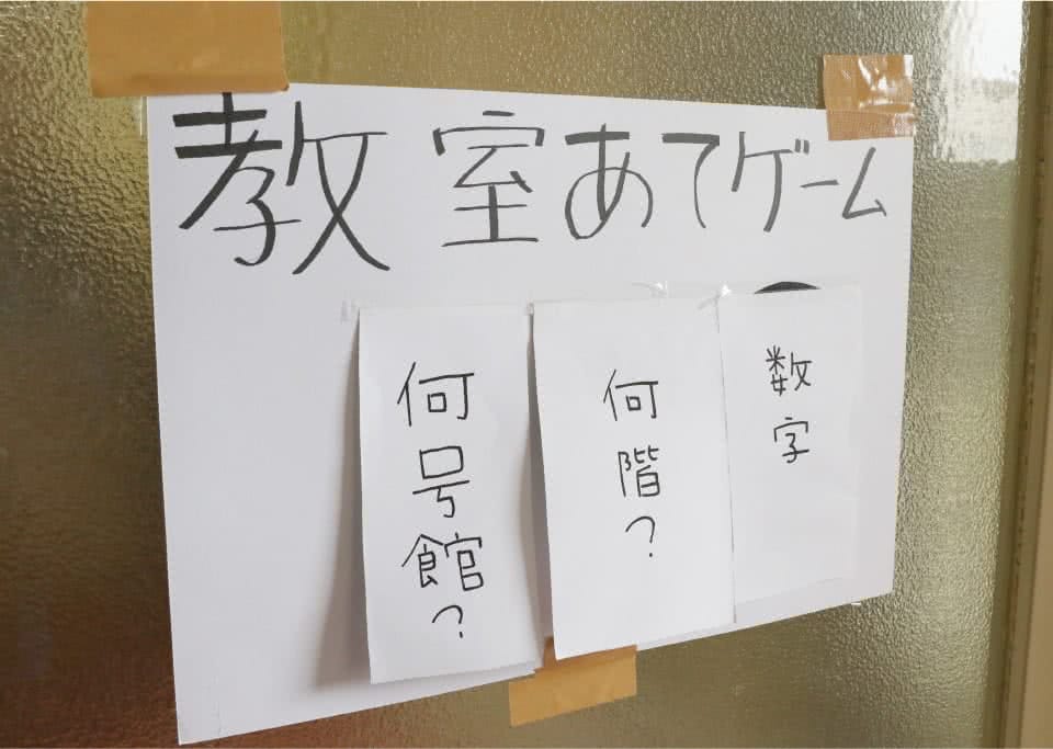 写真:教室あてゲームと書いた画用紙に「何号館?」「何回?」「数字」をと書いた紙が貼られたものをガムテープでガラス戸教室に貼ってあるもの。