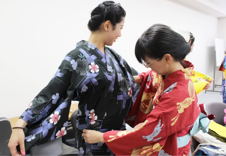 写真:赤い浴衣を着た短大同窓会の先輩に黒い浴衣の着付けをしてもらっている学生