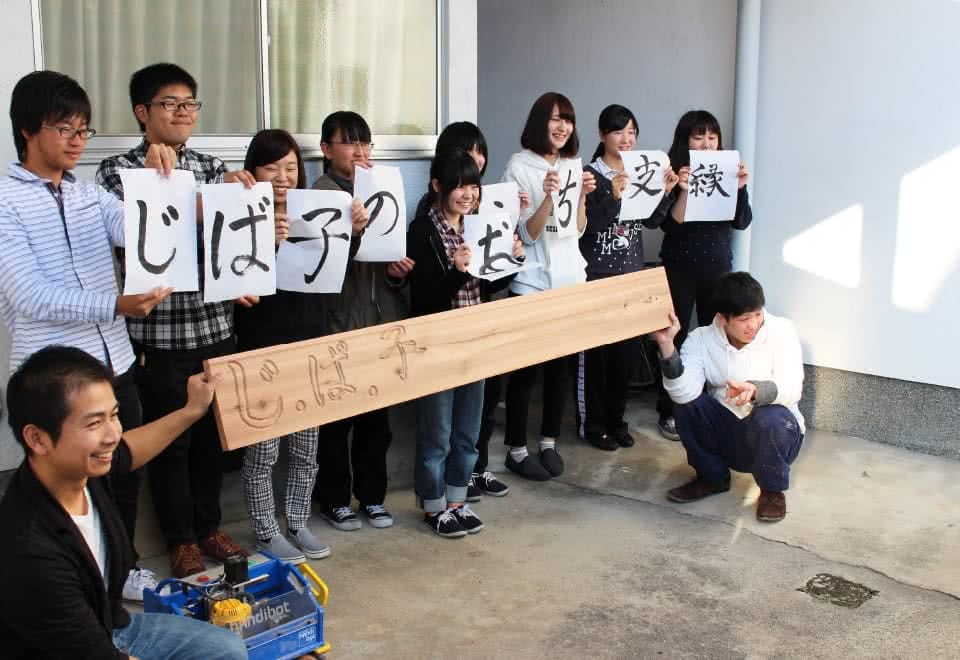 写真:じ・ば・子のおうち支縁オープニングセレモニーにて、看板を持って記念撮影する学生と関係者