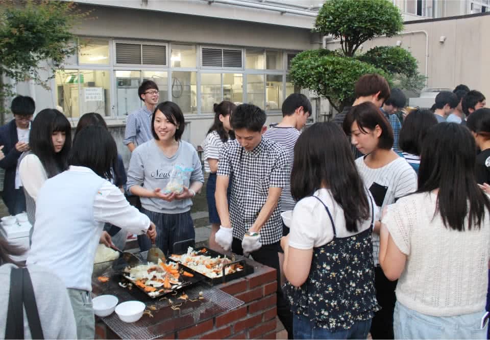 写真:バーベキューコーナーで鉄板で炒め物を作る二人の学生と、それを食べに集まっている学生たち