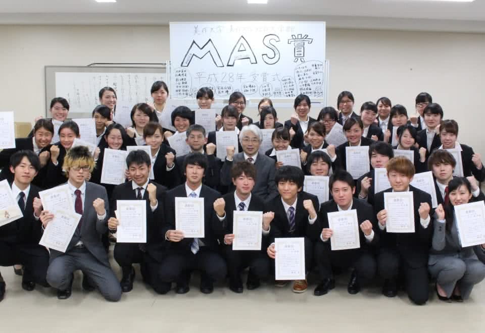 写真:MAS賞と書かれたホワイトボードの前でスーツを着て賞状を持ち、ガッツポーズで集合した学生たちと、その中央にいる学長