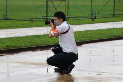 雨が降る中、しゃがんでカメラを構える広報スタッフ