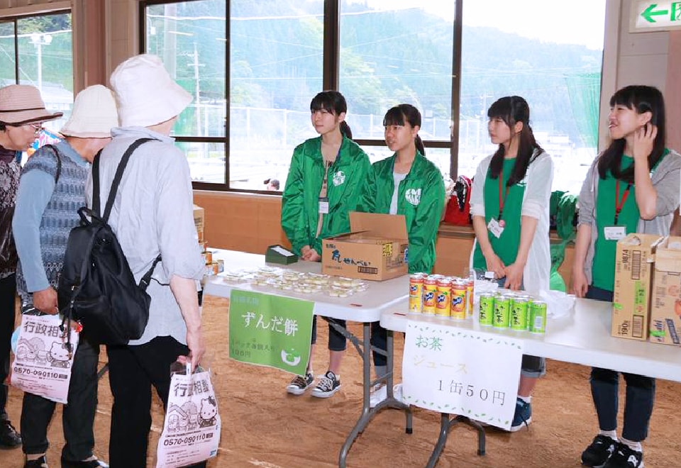 写真：揃いの緑色のジャケットを着て、てずんだ餅やお茶をチャリティマーケットで販売する学生スタッフたち