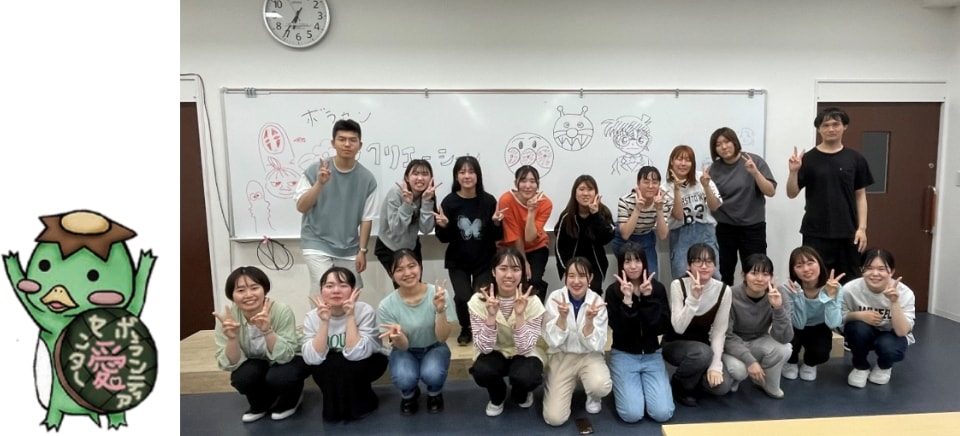ホワイトボードの前でピースサインをして集合写真を撮る学生たちと、その写真の左側に配置されたボランティアセンターのカッパのキャラクター