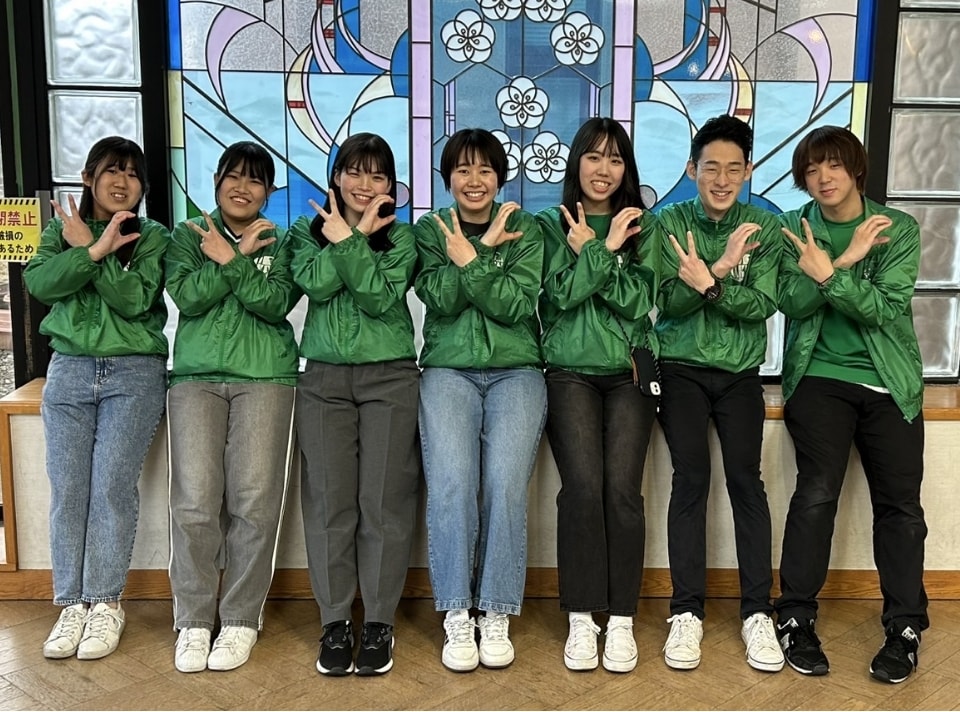 写真:ステンドグラスを背景に、緑色のジャンパー姿でポーズをとる7人の学生
