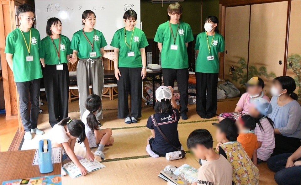 写真:和室で、緑のTシャツを着た学生たちが並んで立ち、座っている子どもたちに説明している様子