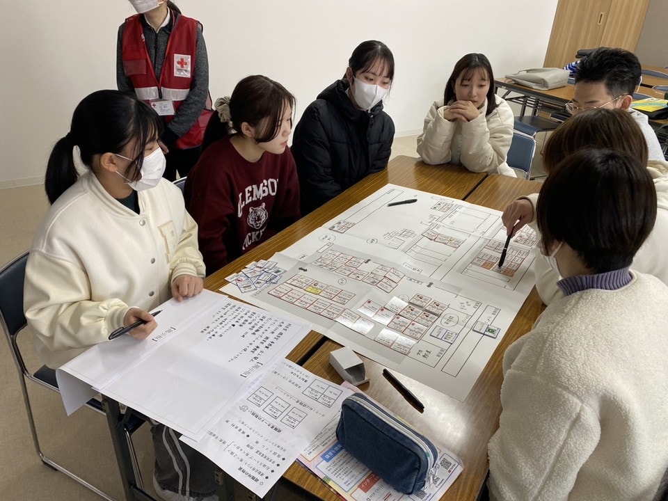 写真:机いっぱいに広げた図面を囲み、真剣な表情で議論する学生たち。後ろには赤十字のスタッフが立って見守っている