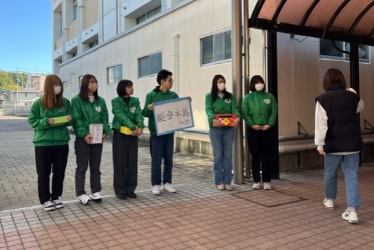 写真:能登半島への募金を呼びかける緑のジャンパーを着た学生グループと、その前を通る学生。校舎前の屋外で撮影