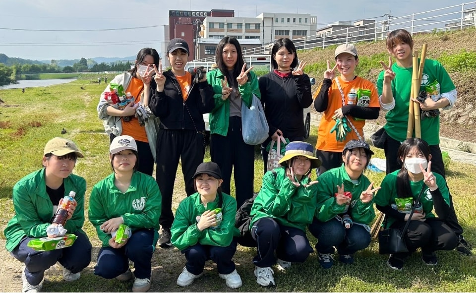 写真:河川敷でごみ拾いを終え、緑のジャンパーやオレンジ色のTシャツ姿で笑顔とピースサインを見せる学生たち