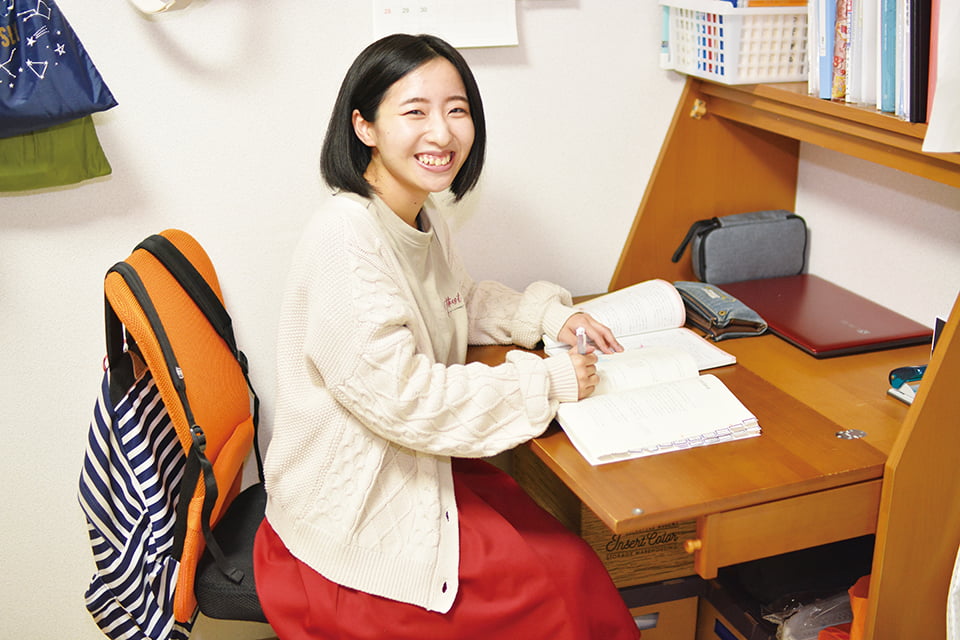 写真:机の上にノートを広げ、右手にペンを持って笑顔を向けるTさん