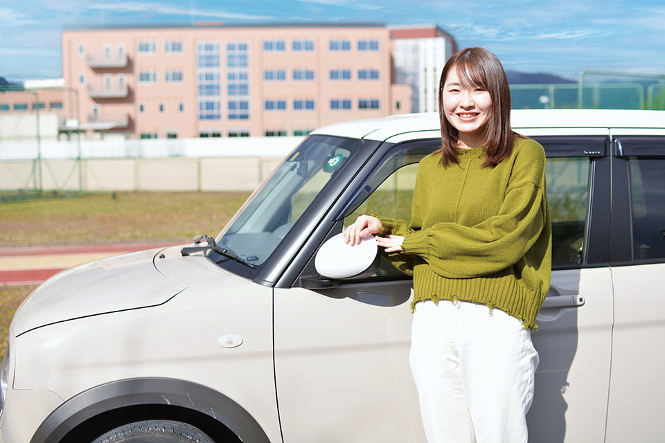 写真:校門前に自動車を駐めて、その横に立ち笑顔を向けるIさん