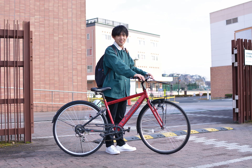 写真：校門前で赤い自転車の横に立つDさん