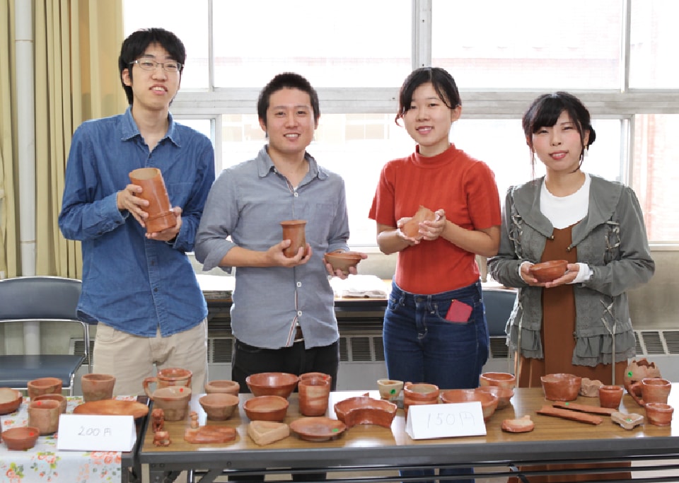 写真：陶芸作品が並べられたテーブルの後ろに立ち、手に作品を持って立つアースワーク部員
