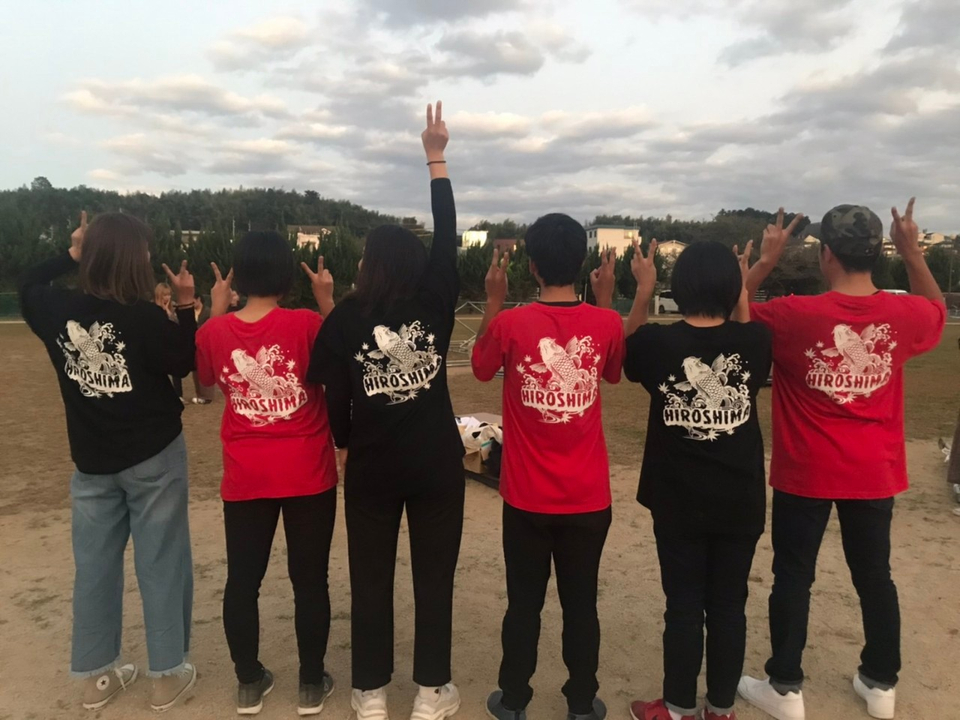 写真：赤と黒のTシャツを着て背中を向けて、ロゴマークを見せながらピースサインをする広島県人会の6人のメンバー