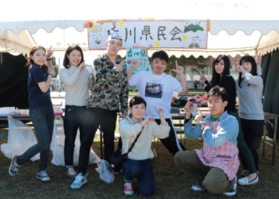 写真：香川県民会の看板を掲げた白梅祭の出店の前で、指を立てたポーズを取る香川県人会のメンバーたち