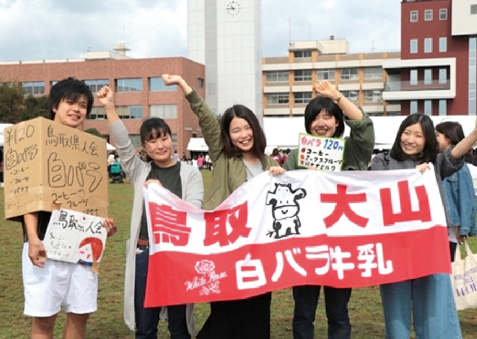 鳥取大山 白バラ牛乳と書かれた垂れ幕を持ち腕を上げる4人の鳥取県人会の女子メンバーと、鳥取県人会 白バラコーヒーと書かれたダンボールをかぶった男子メンバー