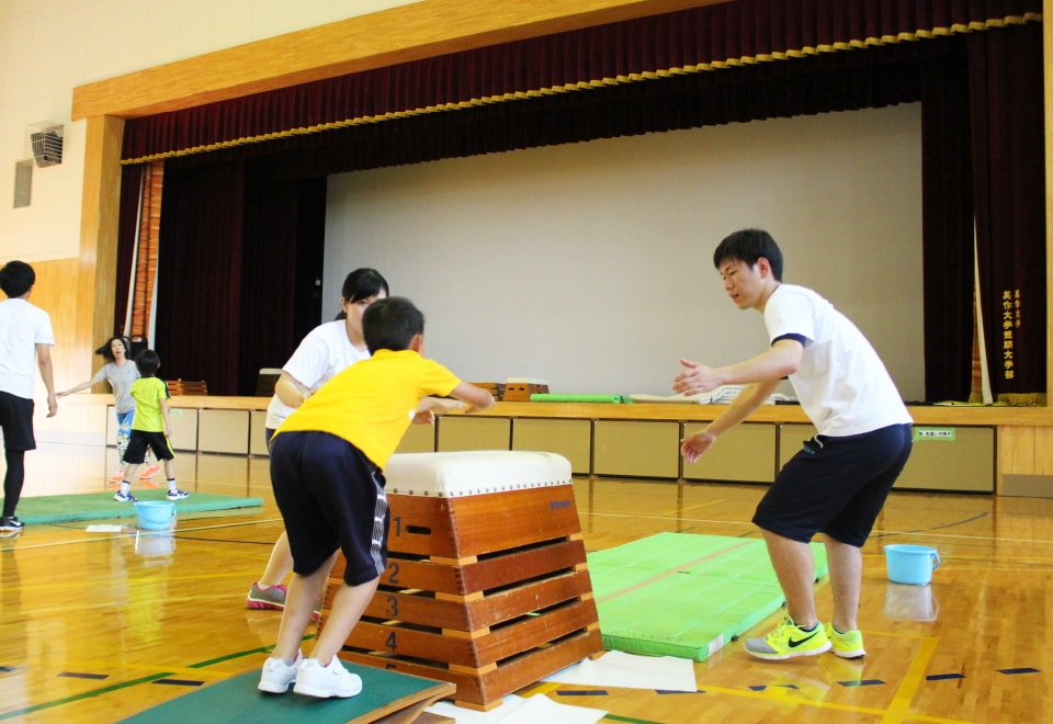 写真：体操教室で跳び箱を飛ぶ小学生と、それをサポートする学生