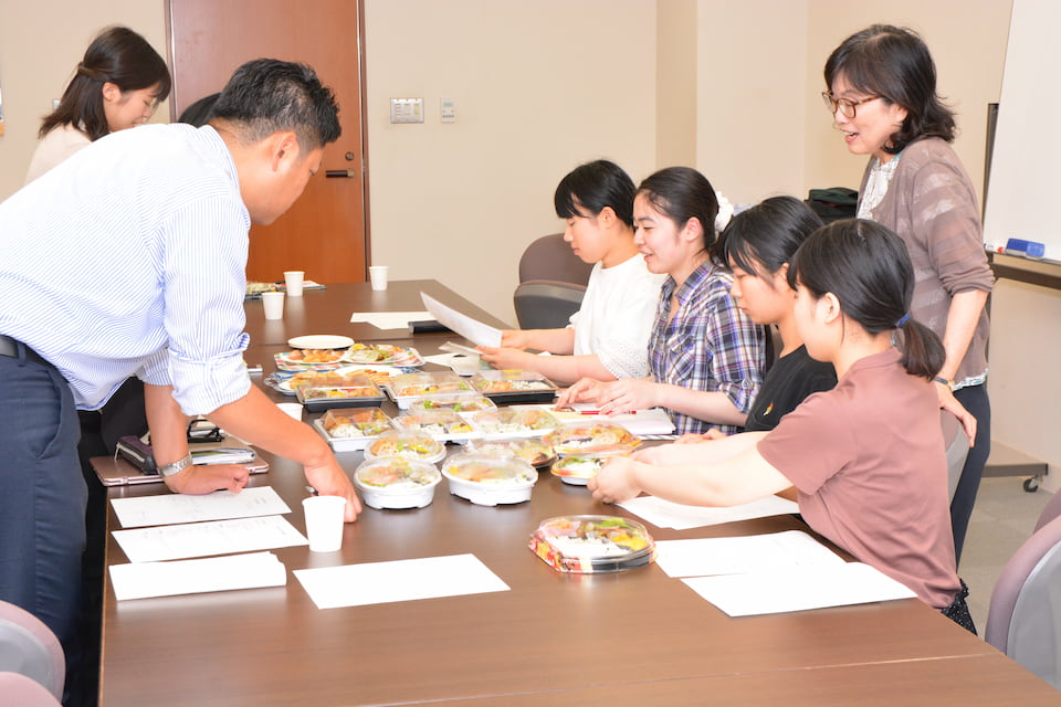 写真：マルイとの弁当開発会議で試作品を試食しながら議論する人見研究室の学生たち・人見准教授とマルイ社員