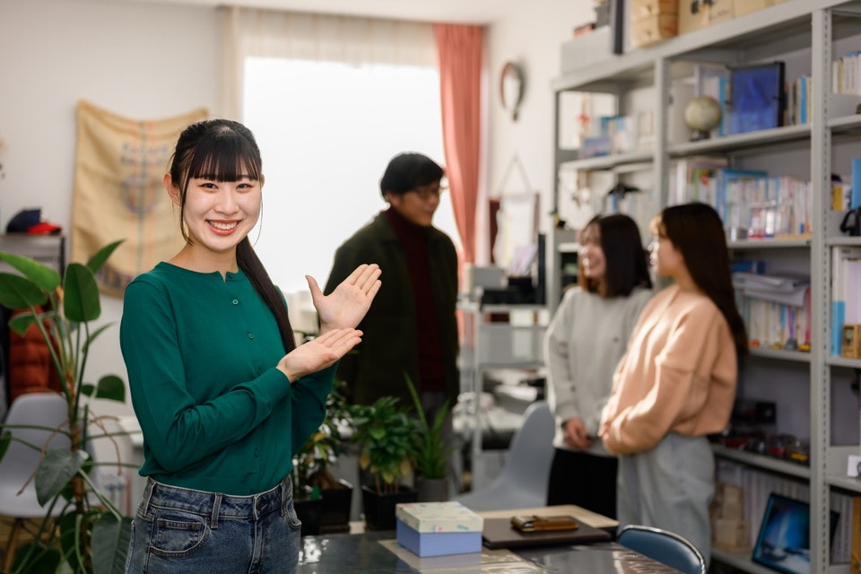 教室で笑顔で手を広げて案内する女性。後ろには本棚と、会話を楽しむ3人の学生がいる