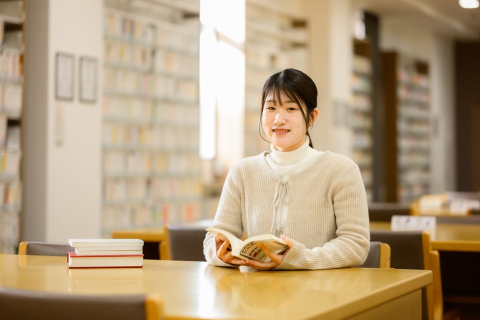 図書館のテーブルで本を手に笑顔を見せる女性学生。隣には数冊の本が積まれている。
