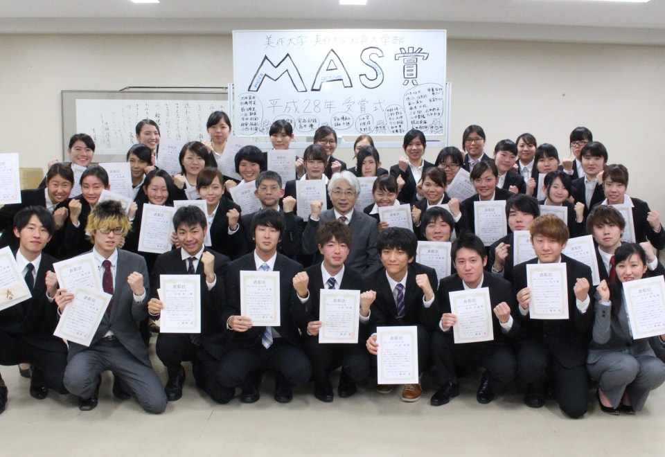 写真：MAS賞と書かれたホワイトボードの前でスーツを着て賞状を持ち、ガッツポーズで集合した学生たちと、その中央にいる学長