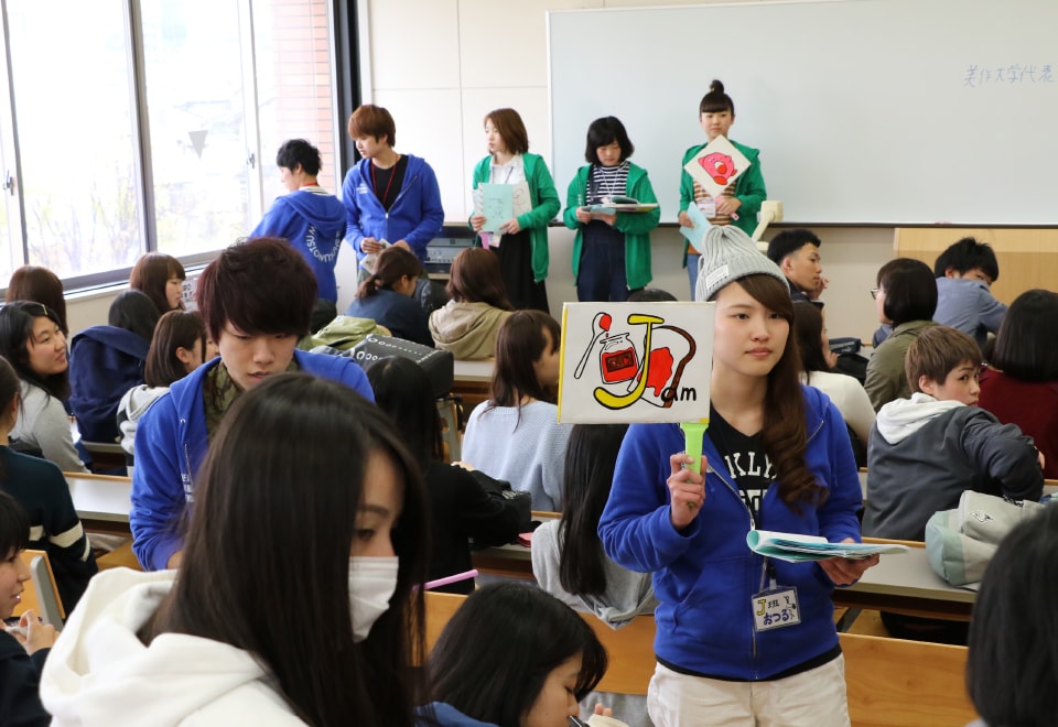写真：教室で着席している新入生と、立っているオリゼミスタッフの学生たち。イラストが描かれた札を持っているオリゼミスタッフの学生もいる。