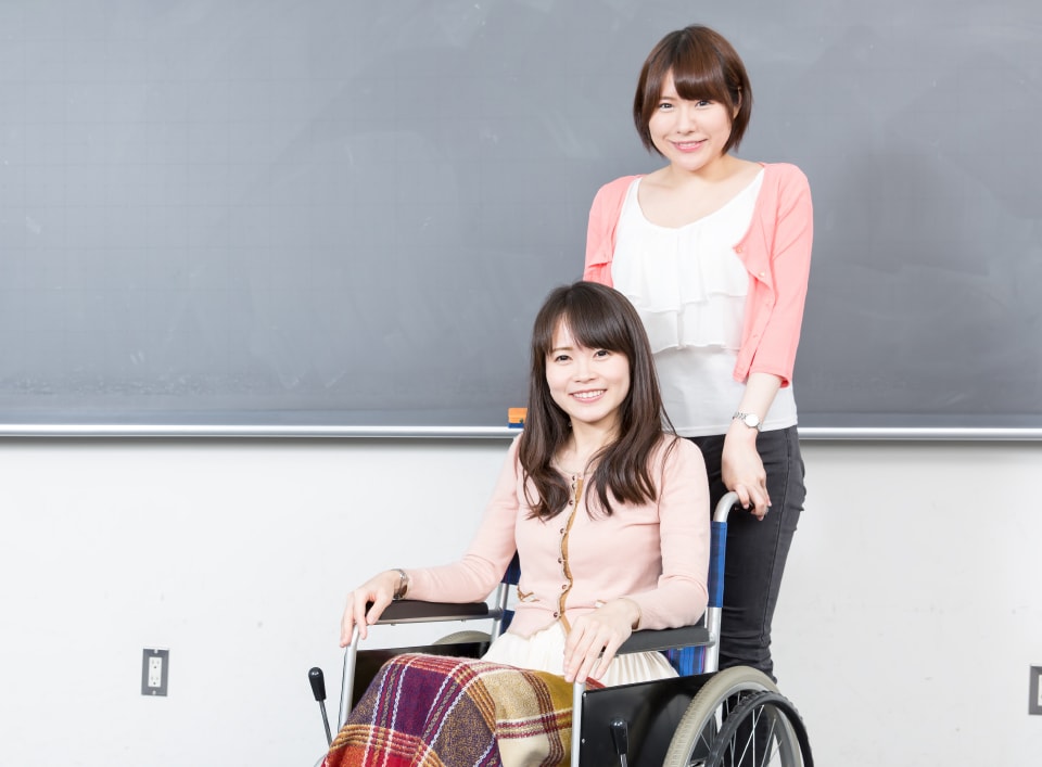 写真：黒板の前で車椅子に座っている女子学生と、その車椅子をもつ女子学生。