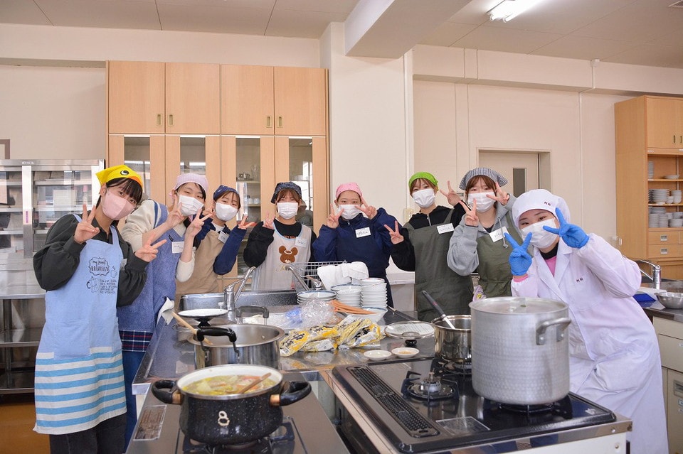 写真:調理実習室でエプロンや帽子、マスクをつけてピースサインをする学生グループ。前には調理中の鍋や食器が並んでいる