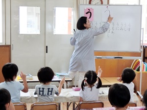 写真：ホワイトボードに「う」を書く平井先生と、席に座って一緒に手を動かしている園児たち