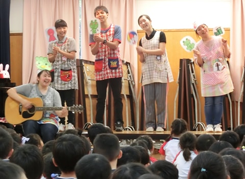 写真：美作大学の生徒たちによる実践演習。園児たちの前で1人がギターを演奏し、4人がイラストが描かれた札を持っている
