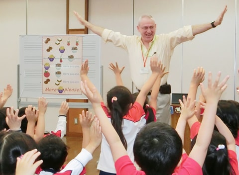 写真：外国人の先生の指導を受ける園児たち。先生に合わせて両手を広げるポースをとっている