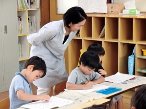 写真：字を書く園児の後ろから指導を行っている平井先生