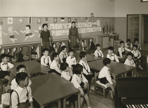 写真：机のある保育室に座っている園児達と壁際に立つ二人の先生