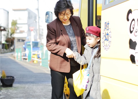 写真：スクールバスから降りてくる女子の園児と手を繋いで補助する先生